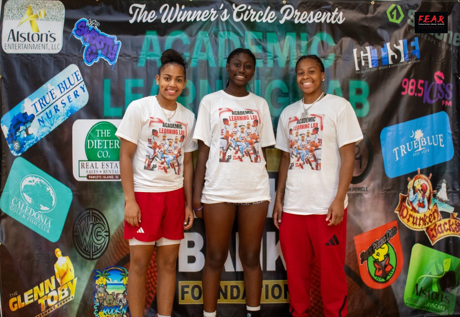Three smiling student athletes in Academic Learning Lab shirts stand before a sponsor banner headed The Winner's Circle Presents Academic Learning Lab, The Book Bank Foundation.