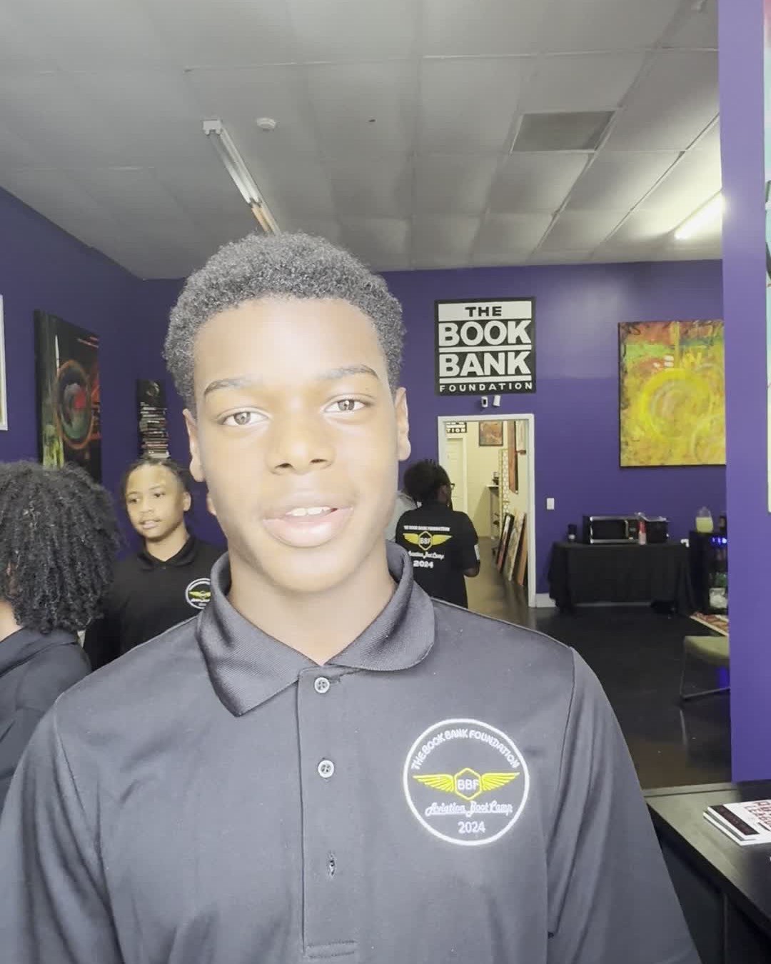 A young cadet in an embroidered Aviation Boot Camp 2024 polo smiles in a Book Bank Foundation space, the foundation's logo on the wall behind him.